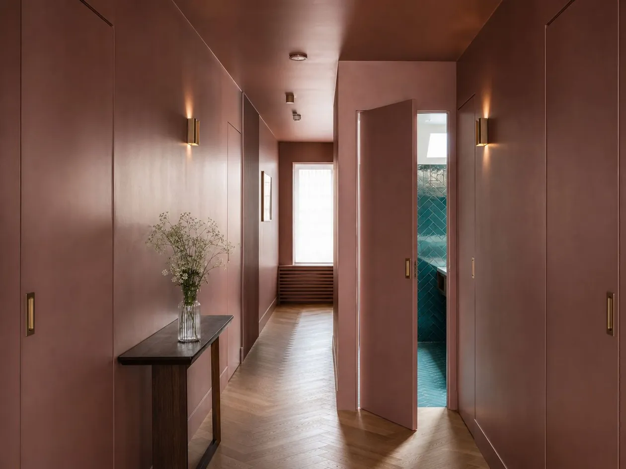 Guest corridor in dusty rose with flush doors, brass finger pulls and a lit room beyond — Boutique Hotel